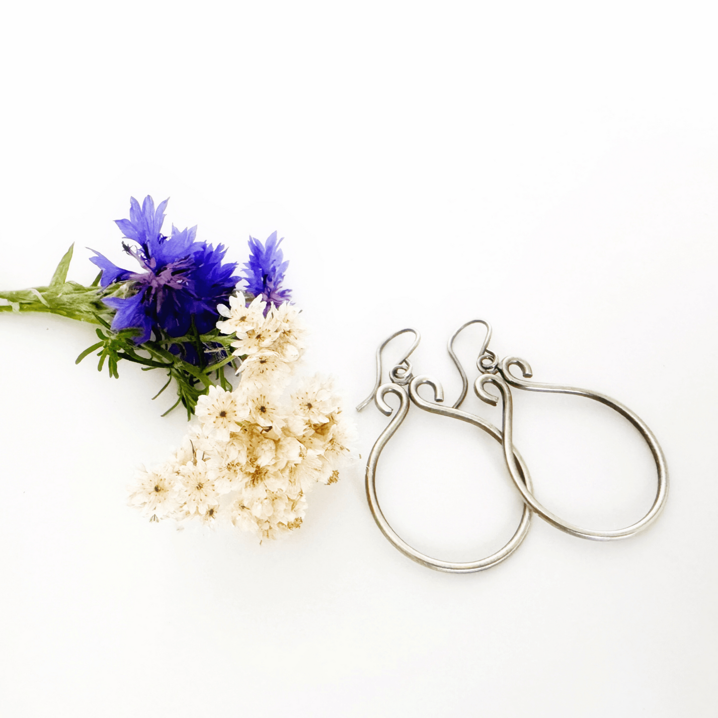 A set of sterling silver hoop earrings on  a white background next to cream and blue flowers. The hoops have been oxidised.